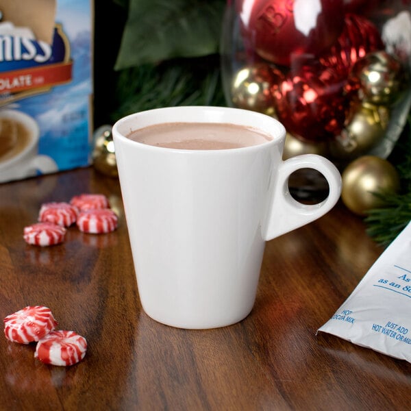 A white mug filled with hot cocoa sits on a wooden surface surrounded by peppermint candies and holiday decorations.