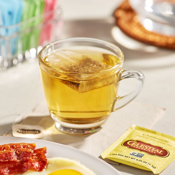 A glass cup of Celestial Seasonings Chamomile Tea with a tea bag on a table with a plate of bacon and eggs.