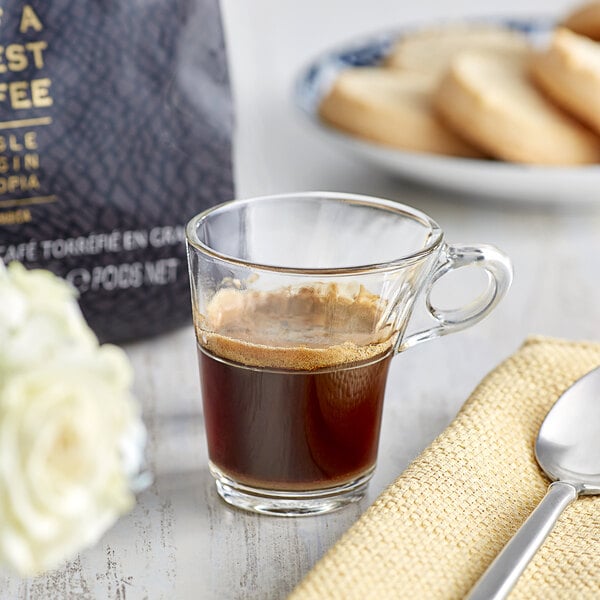 A glass cup of Lavazza espresso on a saucer next to a plate of cookies with a spoon.