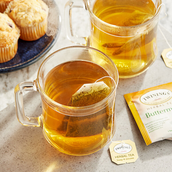 A glass mug of Twinings peppermint and creamy vanilla tea with a tea bag in it on a table with muffins.