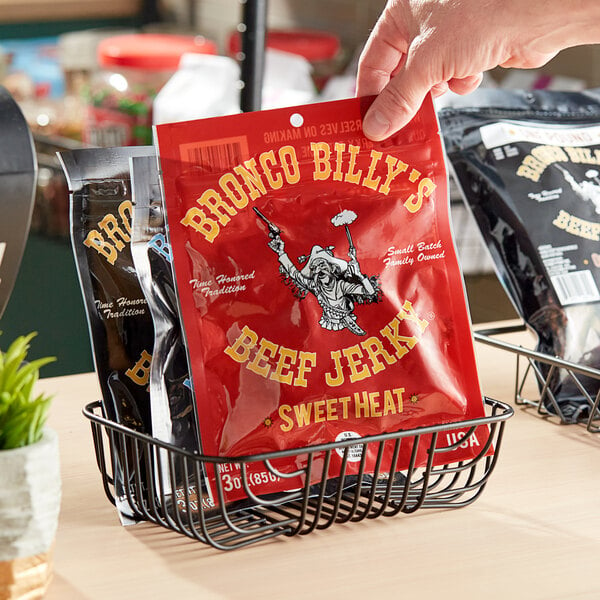 A hand holding a red Bronco Billy's Sweet Heat Beef Jerky packet in a black wire basket on a counter.