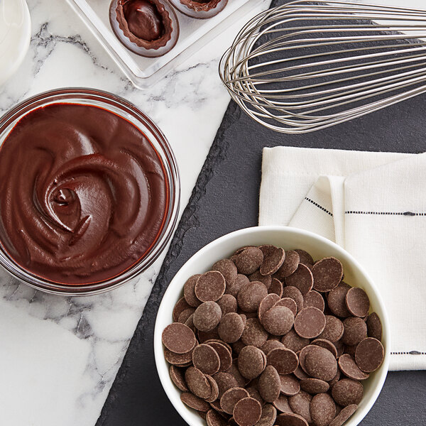 A bowl of Cacao Barry dark chocolate pistoles and a whisk on a black surface.