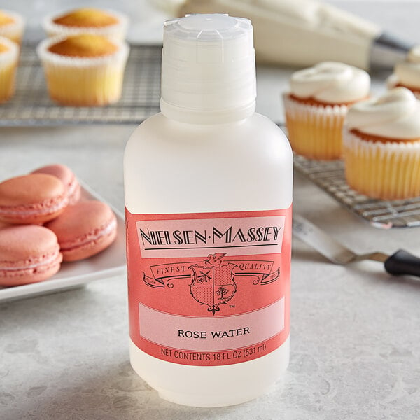 A bottle of Nielsen-Massey rose water on a table with cupcakes and macarons.