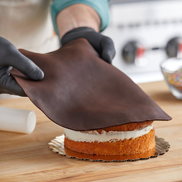 A person wearing gloves rolls a white cylinder of Satin Ice Dark Chocolate Fondant onto a chocolate cake.