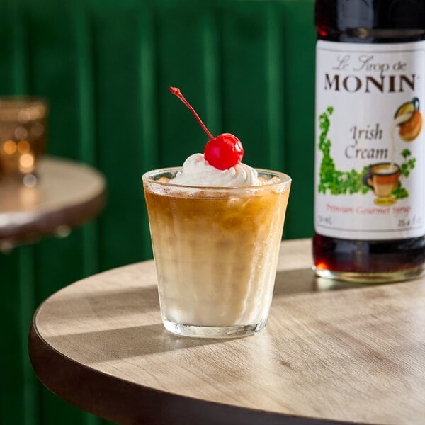 A glass of iced Irish cream-flavored drink topped with whipped cream and a cherry, with a bottle of Monin Irish Cream syrup in the background.