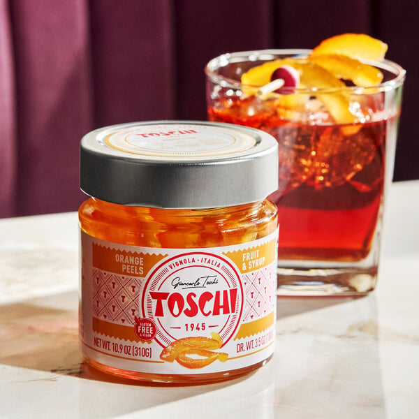 A jar of Toschi orange peels in syrup placed on a table next to a cocktail garnished with orange peel and a cherry.