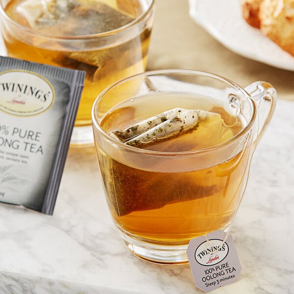 A glass mug and a glass cup of Twinings Oolong tea with tea bags in them on a marble counter.