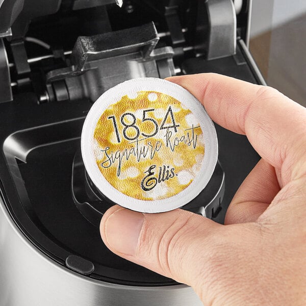 A hand holding a round yellow and white Ellis 1854 Roast Coffee Single Serve Cup container with black text.
