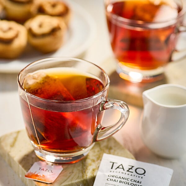 A glass cup of Tazo Organic Chai tea with a tea bag on the table next to a plate of cinnamon rolls.