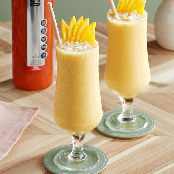 Two glasses of yellow smoothies with pineapple slices and yellow liquid in a SHOTT Mango Real Fruit Flavoring Syrup container.