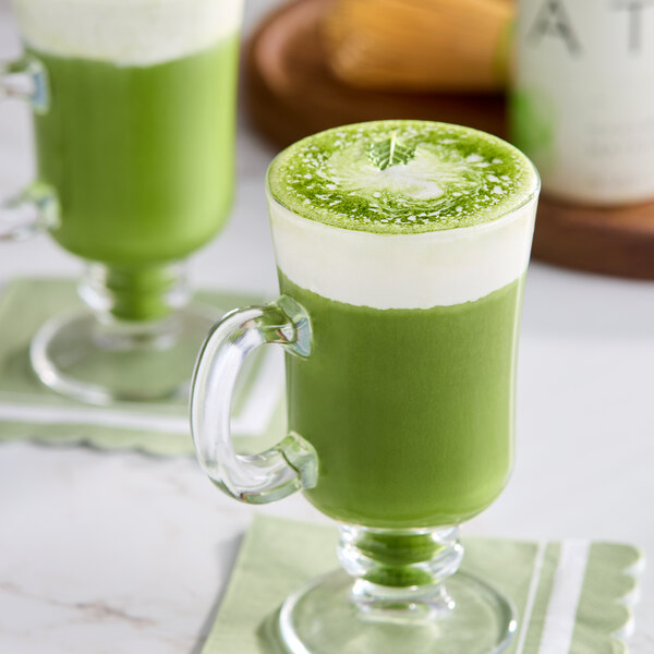 A glass mug filled with a green matcha latte topped with a layer of frothy milk.