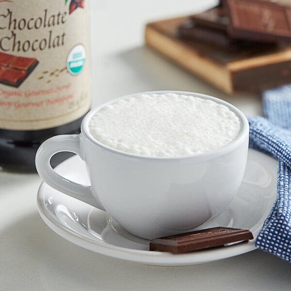 A white cup of hot chocolate with foam and a bottle of Monin Organic Chocolate Flavoring syrup.