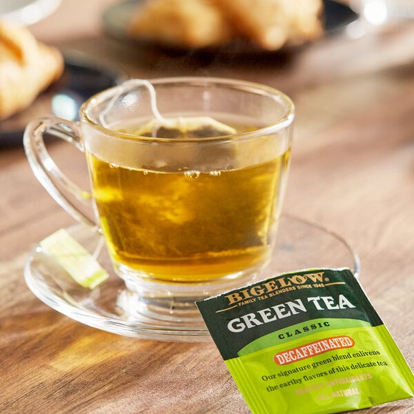 A glass cup of Bigelow decaffeinated green tea with a Bigelow green tea bag on a saucer.