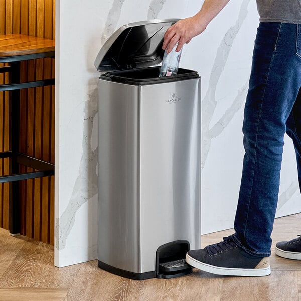 A man using a Lancaster Table & Seating rectangular decorative step-on trash can to throw away a bottle.