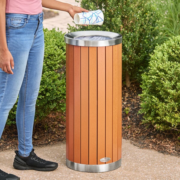 A round outdoor trash can with a wood composite exterior and a stainless steel open lid.