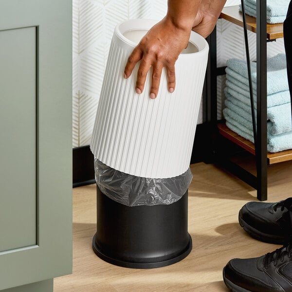 A white round decorative wastebasket with a black base and a plastic liner being placed inside.