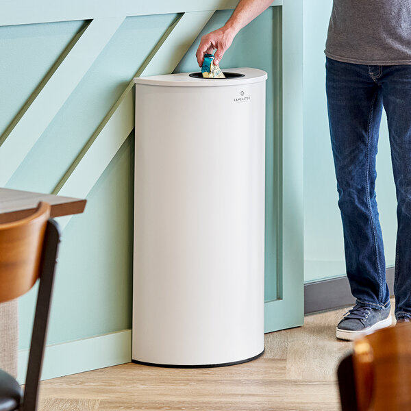 A person putting a piece of paper in a white half round Lancaster Table & Seating decorative trash can.