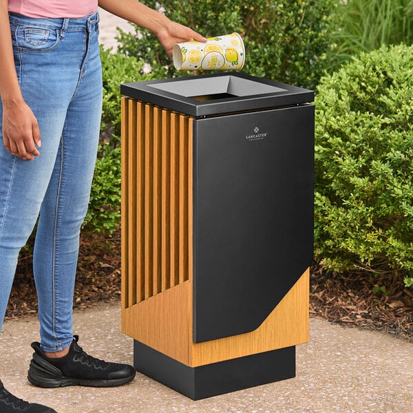 A square decorative trash can made of steel and wood with a modern design, placed outdoors.