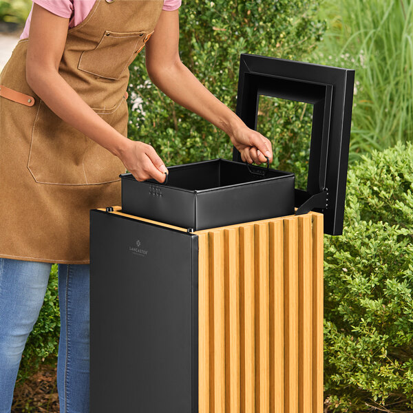 A square decorative trash can with a steel and wood design, shown with its lid open and an inner bin being removed.