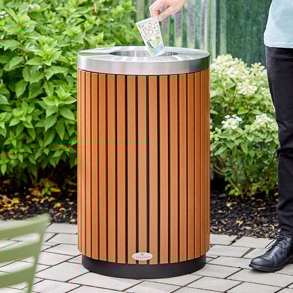 A round outdoor trash can with a wood composite slat design and a stainless steel open lid.