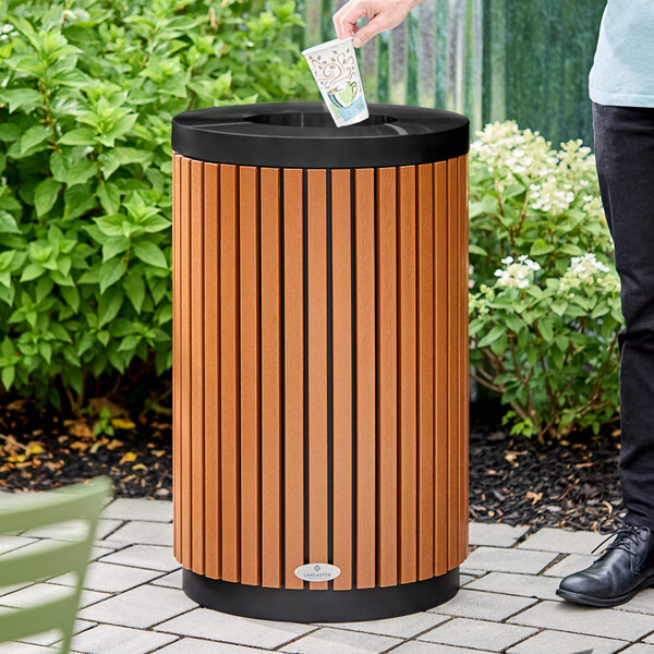 A person is placing a paper cup into a round outdoor trash can with vertical wood-like slats and a black open lid, situated on a paved area with greenery in the background.