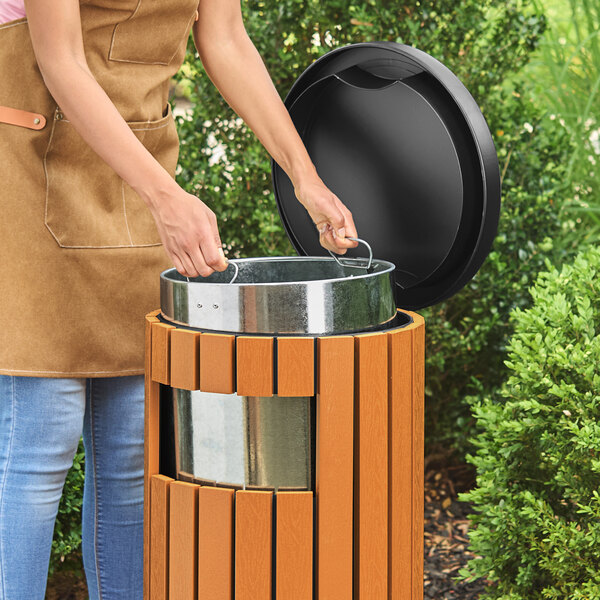 A round outdoor trash can with a wood composite exterior, steel inner bin, and a side opening lid.