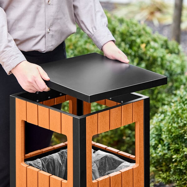 A man holding a Lancaster Table & Seating square black and brown decorative trash can with covered top.