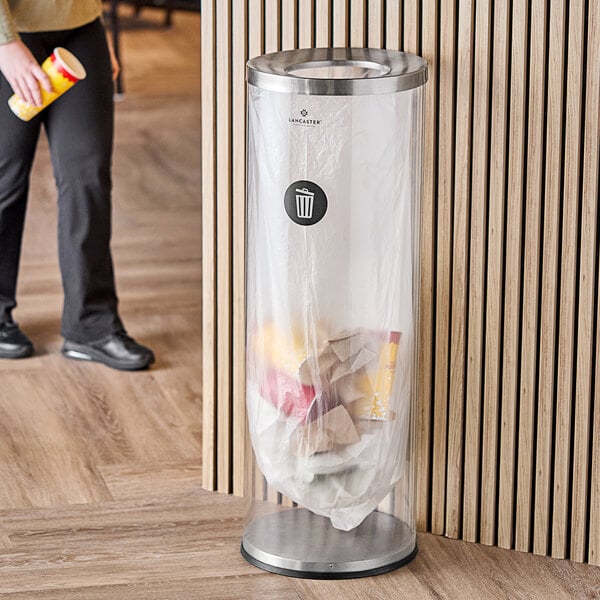 A person standing next to a Lancaster Table & Seating clear plastic decorative trash can with a stainless steel top.