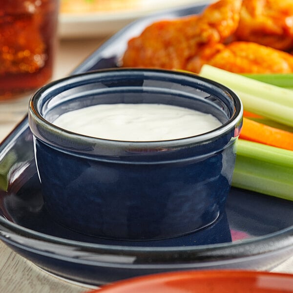 A plate with a blue Acopa Azora ramekin filled with white liquid next to celery sticks.