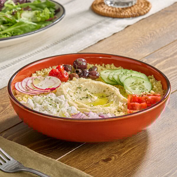 An orange porcelain coupe low bowl filled with hummus, couscous, sliced cucumbers, radishes, tomatoes, olives, feta cheese, and red onions.