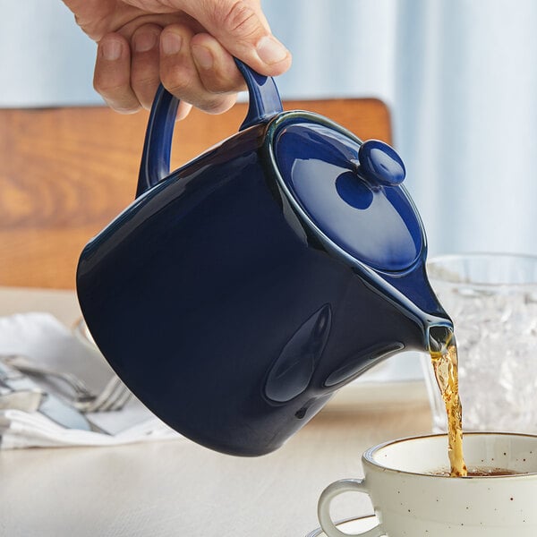 A hand pouring tea into a blue Acopa Keystone teapot.