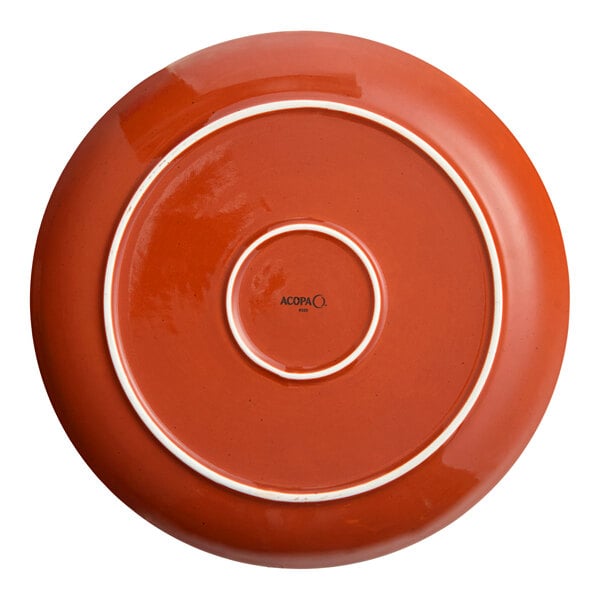 An Acopa Keystone Sedona Orange porcelain coupe plate with a white rim on a red surface.