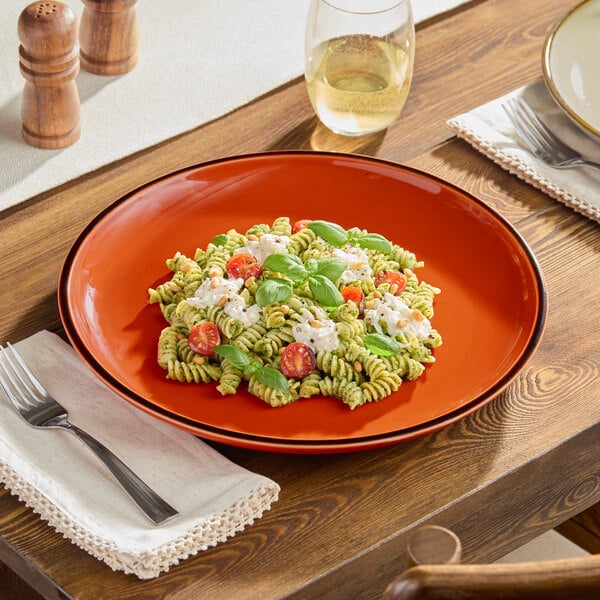A plate of pasta with tomatoes and basil on an Acopa Keystone Sedona Orange porcelain coupe plate on a wooden table.
