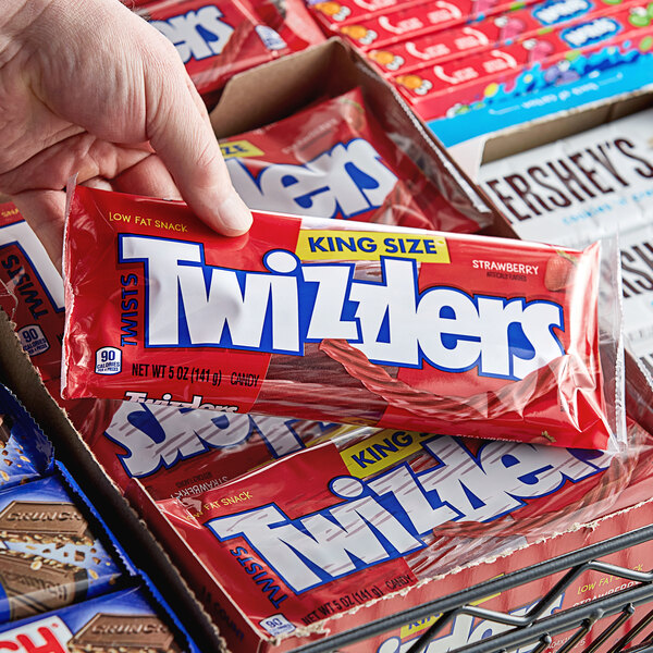 A hand holding a red box of TWIZZLERS on a table in a convenience store.