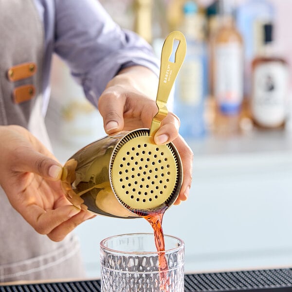 A person using a Barfly gold-plated Hawthorne strainer to pour a drink from a shaker.