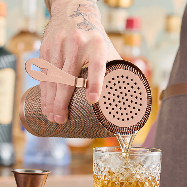A man using a Barfly copper-plated Hawthorne strainer to pour a drink into a glass.