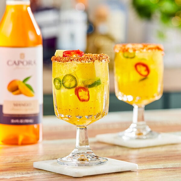 A glass of yellow mango-flavored beverage with ice, chili slices, and a spiced rim, next to a bottle of Capora Mango Flavoring Syrup.