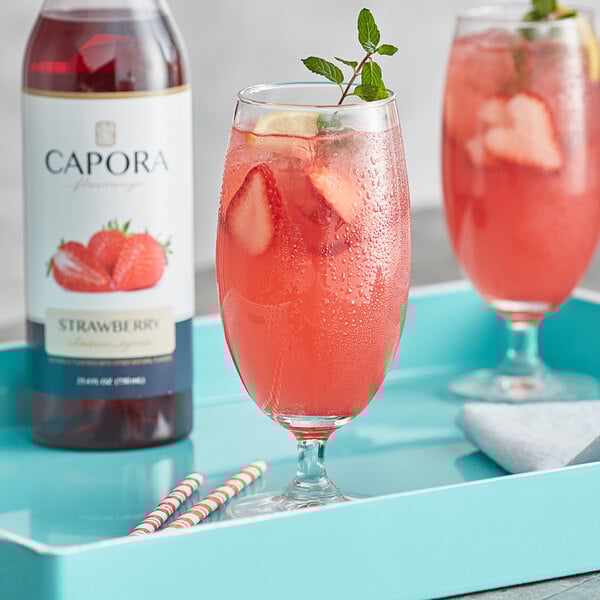 A glass of red liquid flavored with Capora Strawberry Syrup next to a white bottle of Capora Strawberry Flavoring Syrup.