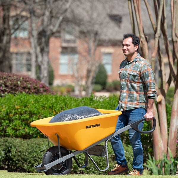 A man pushing a yellow True Temper wheelbarrow with black plastic bag in it.