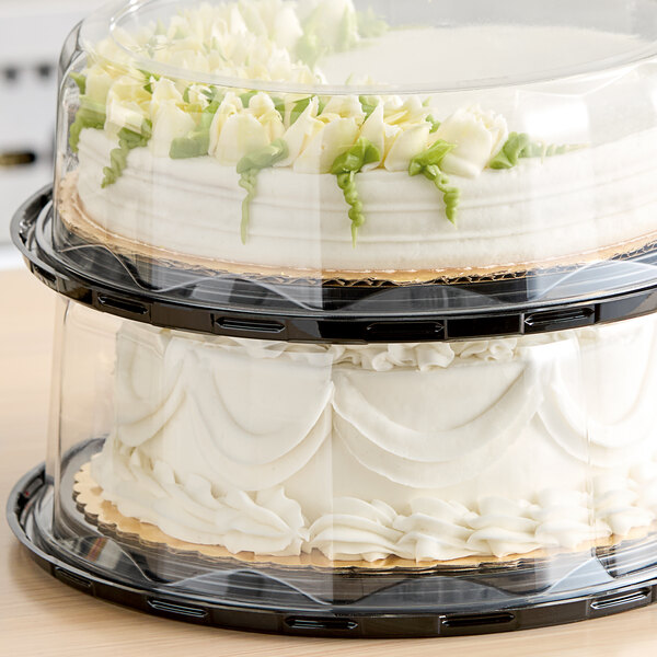 Two Baker's Lane cake trays with a white cake and green leaf decorations, one with a clear plastic scalloped cover.