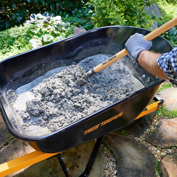A black steel wheelbarrow with a flat-free tire being used to mix concrete outdoors.