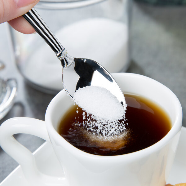A hand holding a Oneida Unity stainless steel demitasse spoon pouring sugar into a cup of coffee.