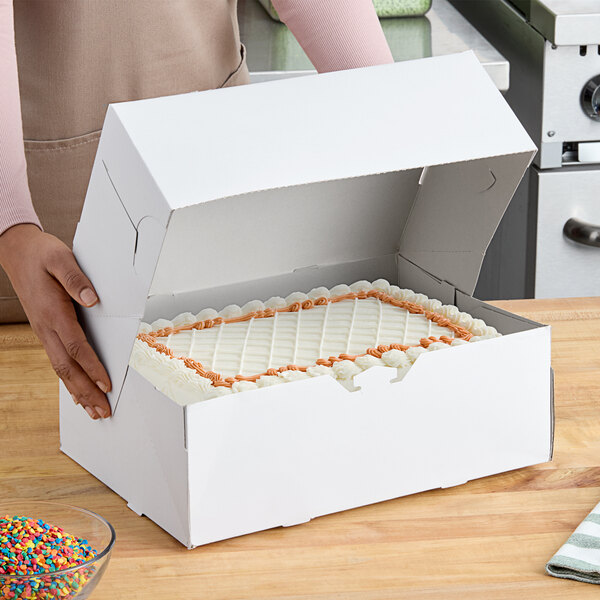 A person holding a Baker's Lane white cake box with a cake inside.