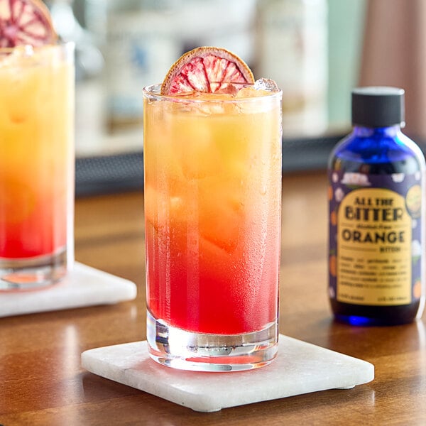 A glass of orange drink on a coaster with a bottle of All The Bitter Non-Alcoholic Orange Bitters.