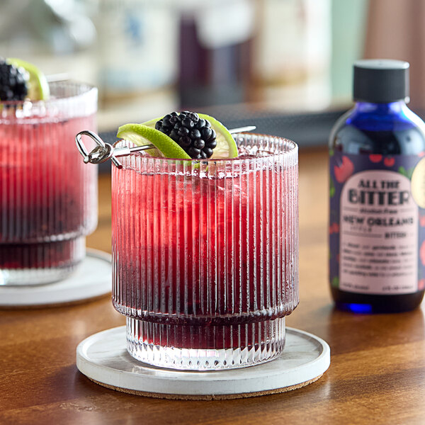 A glass of red drink with a straw and fruit next to a bottle of All The Bitter New Orleans-Style Bitters.
