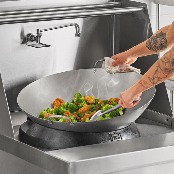 A large carbon steel Cantonese wok being used to stir-fry vegetables and chicken in a commercial kitchen setting.