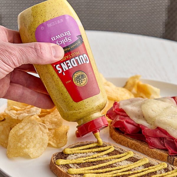 A hand holding a Gulden's Spicy Brown Mustard squeeze bottle over a sandwich.