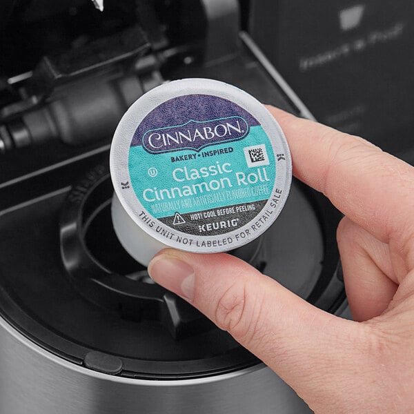 A hand holding a round blue and white container of Cinnabon Classic Cinnamon Roll coffee.