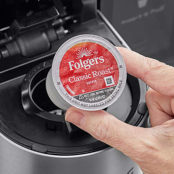 A hand holding a red and white round container of Folgers Classic Roast Coffee.