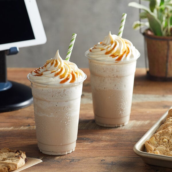 Two cups of Big Train Caramel Latte Blended Ice Coffee on a table with cookies and straws.